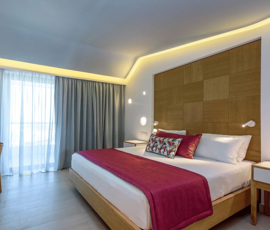 Lyttos Mare Superior Room featuring a large bed with red and white pillows, a wooden headboard, and ambient lighting, with a view outside the window.