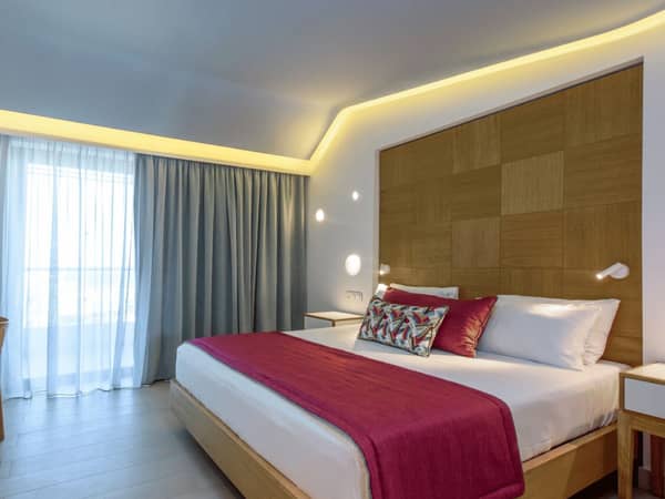 Lyttos Mare Superior Room featuring a large bed with red and white pillows, a wooden headboard, and ambient lighting, with a view outside the window.
