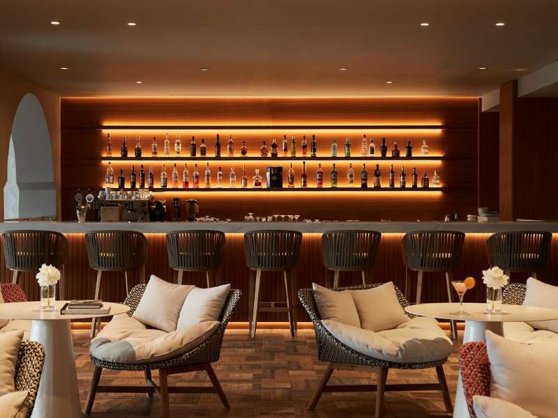 Sophisticated bar in Marbella Gallery, with wooden bar counter, high stools, and ambient lighting, featuring an array of spirits on shelves.
