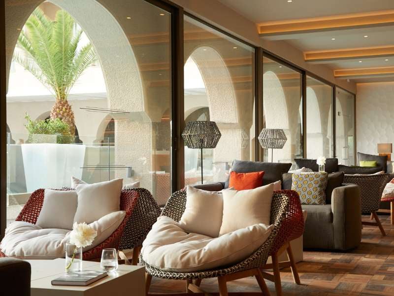 Modern lounge in a Marbella gallery with wicker armchairs, plush cushions, glass wall view of a palm tree, and elegant interior design.