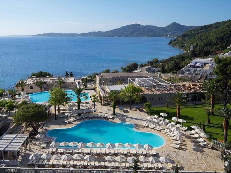 Aerial view of Marbella Gallery resort with two heart-shaped swimming pools, surrounded by sun loungers and palm trees, overlooking the sea and mountains.
