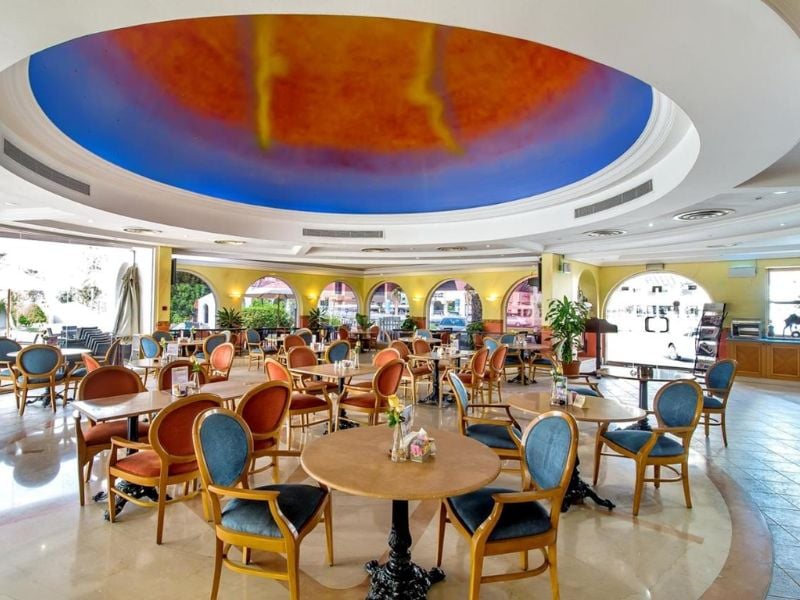 Interior of Marbella Gallery Restaurant with wooden chairs, circular tables, and a ceiling mural in orange and blue hues.