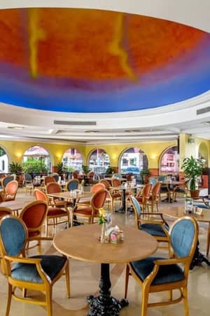 Interior of Marbella Gallery Restaurant with wooden chairs, circular tables, and a ceiling mural in orange and blue hues.