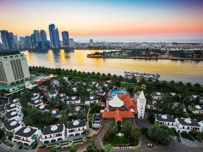 Aerial sunset view of Marbella Gallery, featuring luxury villas with palm trees, overlooking waterfront and high-rise buildings in the distance.