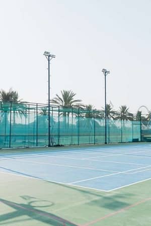 Outdoor tennis court in Marbella with green surface, surrounded by high fence and palm trees, featuring floodlights for evening games.
