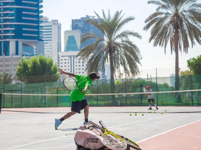 Two young tennis players practising on a court in Marbella, with one hitting a volley and palm trees and skyscrapers in the background.