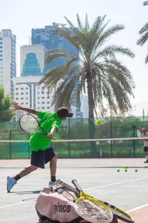 Two young tennis players practising on a court in Marbella, with one hitting a volley and palm trees and skyscrapers in the background.