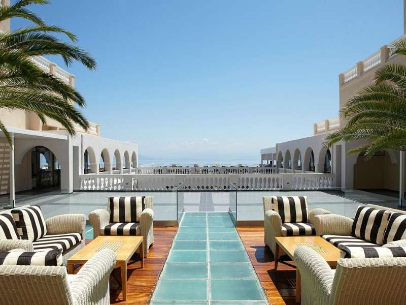 Veranda at Marbella Gallery with elegant striped furniture and palm trees, overlooking a sea view with grand arches.