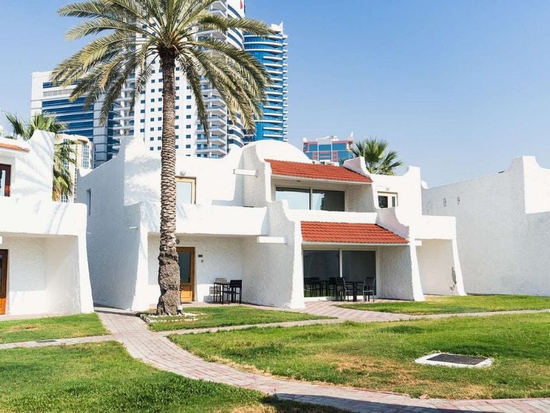 White-washed villas at Marbella Gallery with green lawns, a palm tree, and skyscrapers in the background.