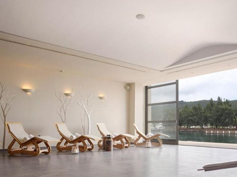 Indoor lounge of Meliton Gallery with wooden reclining chairs and a large window showing a lake and greenery.