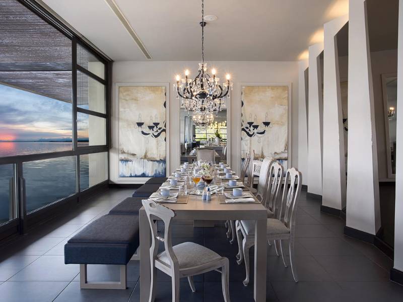 Dining room at Meliton Gallery Restaurant, with a long table set, chandeliers, large windows showing a sunset view, and modern art on the walls.