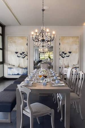 Dining room at Meliton Gallery Restaurant, with a long table set, chandeliers, large windows showing a sunset view, and modern art on the walls.