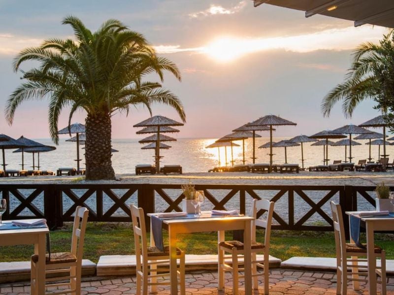 Seafront dining area at Meliton Gallery with wooden tables, chairs, straw parasols, and a sunset over the ocean.