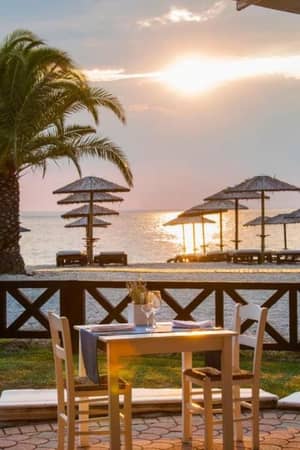 Seafront dining area at Meliton Gallery with wooden tables, chairs, straw parasols, and a sunset over the ocean.