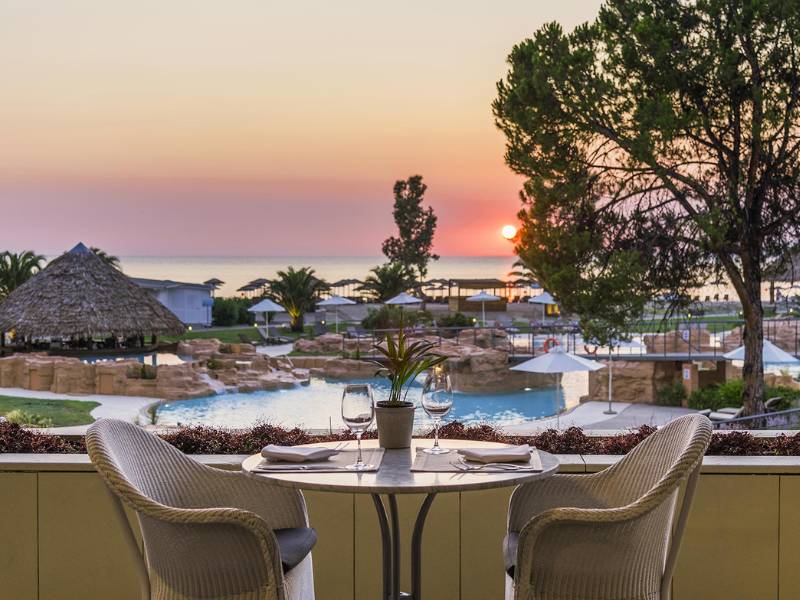 Dinner table set for two overlooking a pool at Meliton Gallery, with a sunset view over the sea and surrounding palm trees.