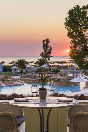 Dinner table set for two overlooking a pool at Meliton Gallery, with a sunset view over the sea and surrounding palm trees.