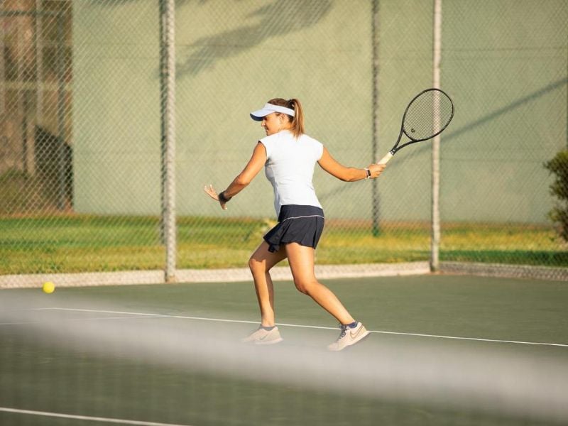 Female tennis player prepares for a forehand shot on an outdoor court, wearing a visor and sports attire, with sunlight highlighting the scene.
