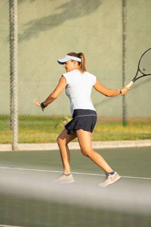 Female tennis player prepares for a forehand shot on an outdoor court, wearing a visor and sports attire, with sunlight highlighting the scene.