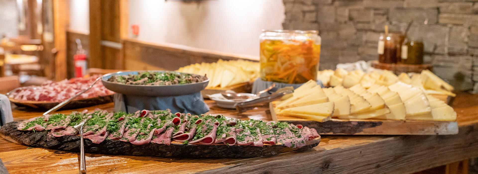 Gourmet food spread at Mira Alagna Mountain Resort & Spa, showing cured meats with herbs, sliced cheeses, and pickled vegetables against a stone wall.