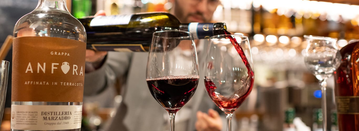 Bartender pouring red wine into glasses in a cosy bar at Mira Alagna Mountain Resort & Spa, with Anfora Grappa bottle visible.