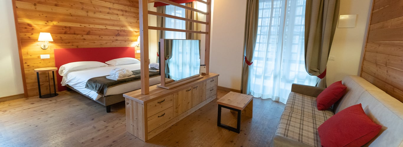 Room interior at Mira Alagna Mountain Resort & Spa featuring a double bed, wooden partition, TV, plaid sofa, and large windows.