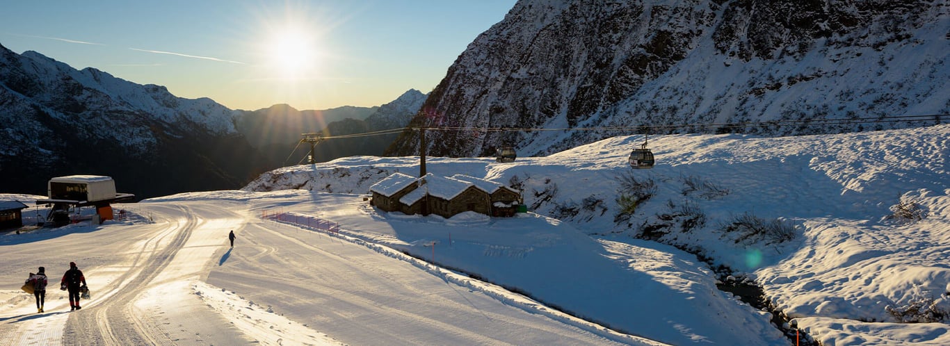 Skiers on snow-covered Mira Alagna Mountain Resort slopes with mountains and a sunset in the background.