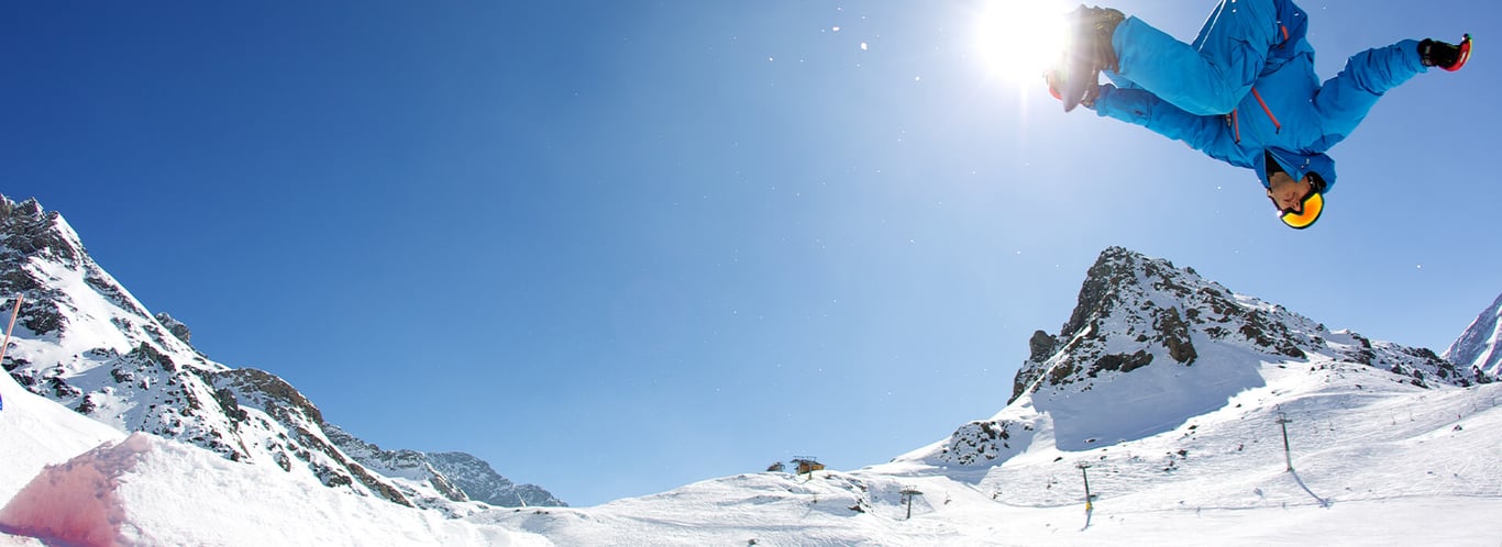Ski jumper in vibrant blue gear soaring over snow-covered slopes at Mira Alagna Mountain Resort & Spa with mountains in the background.