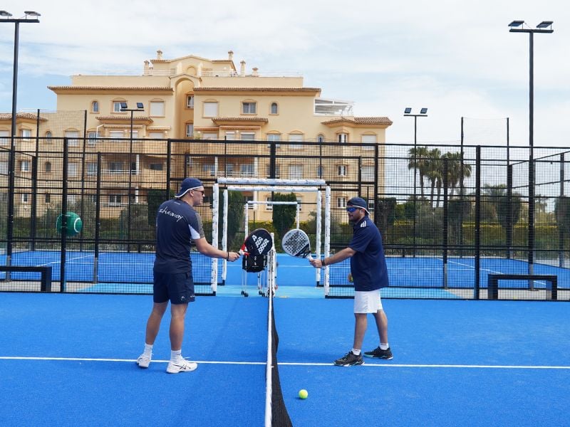 Two coaches on a blue padel court at Oliva Nova Beach Resort, holding racquets and having a discussion with luxury apartments in the background.