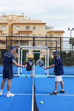 Two coaches on a blue padel court at Oliva Nova Beach Resort, holding racquets and having a discussion with luxury apartments in the background.