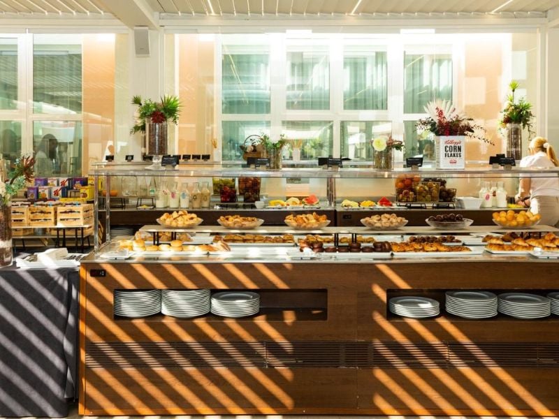 Buffet at Oliva Nova Padel with pastries, fruits, and breakfast items in a bright dining area with glass windows and potted plants.