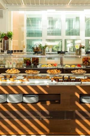 Buffet at Oliva Nova Padel with pastries, fruits, and breakfast items in a bright dining area with glass windows and potted plants.