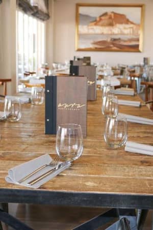 Oliva Nova dining area with a long wooden table, wine glasses, napkins, and contemporary chairs, framed by a large painting.