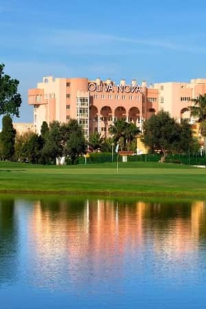 Oliva Nova Resort exterior with a golf course, palm trees, and a water feature under a blue sky.