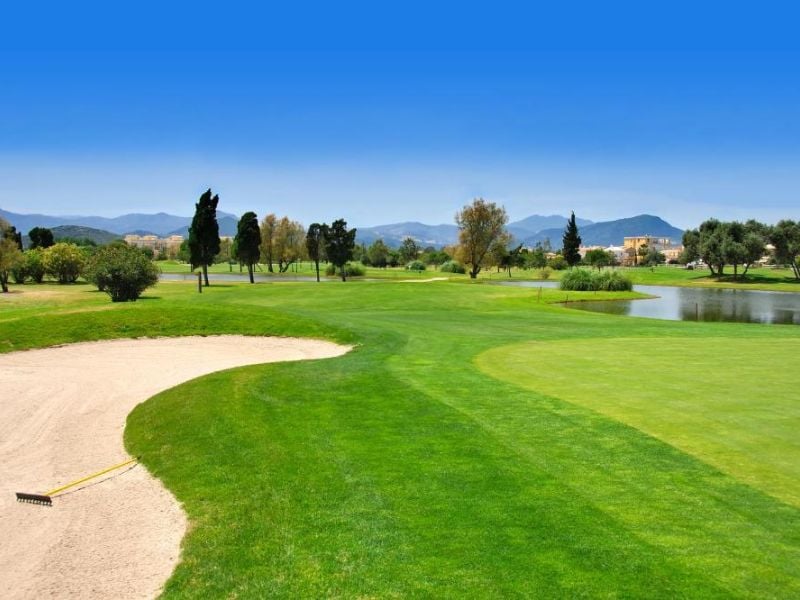 Golf course at Oliva Nova, featuring a sand bunker, fairways, a water hazard, and mountains in the backdrop.