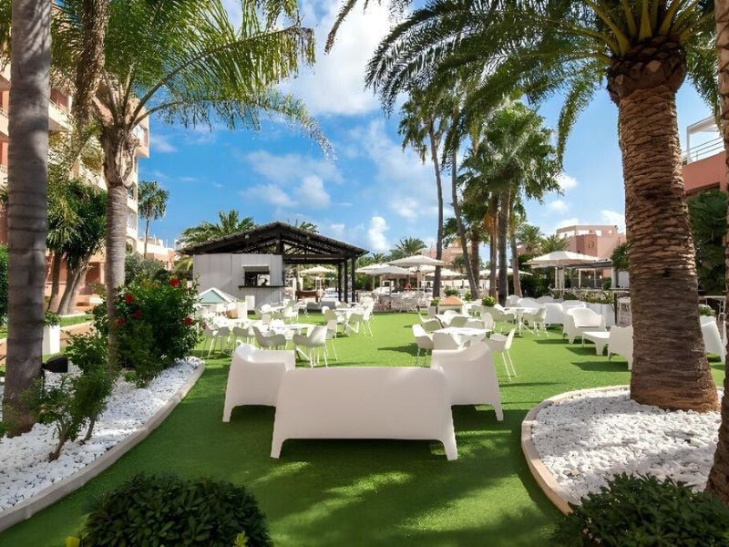 Outdoor lounge at Oliva Nova with white furniture, palm trees, and green artificial turf.