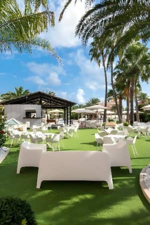 Outdoor lounge at Oliva Nova with white furniture, palm trees, and green artificial turf.