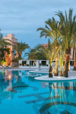 Outdoor pool at Oliva Nova Resort with palm trees, modern cabanas, and dusk lighting reflecting on the water surface.