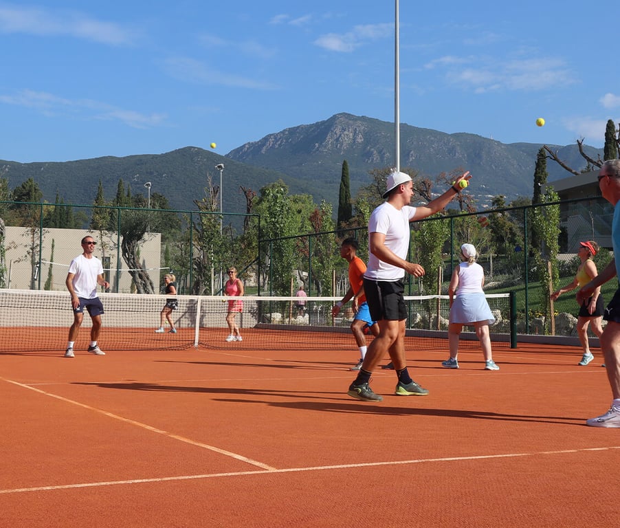 People playing tennis on a clay court with mountains and greenery in the background.