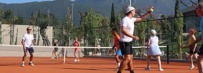 People playing tennis on a clay court with mountains and greenery in the background.