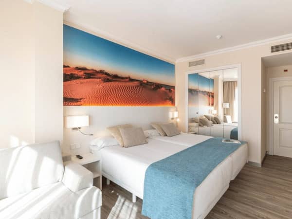 Oliva Nova Golf Resort room with two double beds, beach mural, grey sofa, and modern decor.