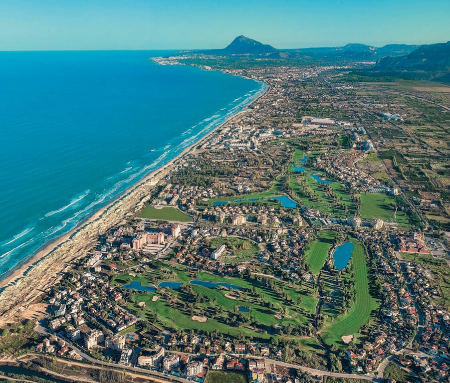 Aerial view of Oliva Nova highlighting golf courses, residential areas, and the Mediterranean coastline with mountains in the background.