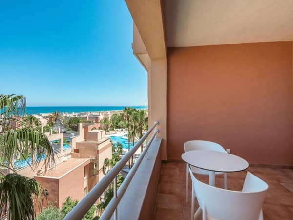 Balcony of an Oliva Nova sea view room with white table and chairs, overlooking the resort, palm trees, and ocean.