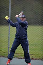 Ollie Kirk in a navy tracksuit poised to hit a tennis ball on an outdoor court.