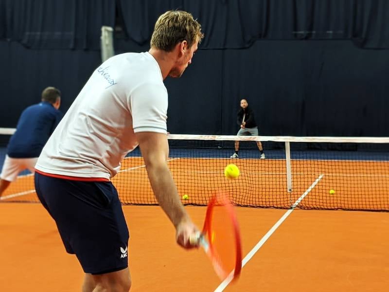 Tennis coaching session at PW Nottingham, featuring a coach preparing to hit a tennis ball with a red racket on indoor courts.