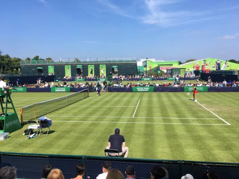 Outdoor tennis match at Nottingham Tennis Centre, with grassy court, players in action, and audience in stands on a sunny day.