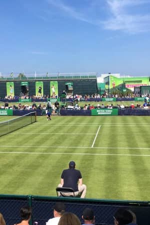 Outdoor tennis match at Nottingham Tennis Centre, with grassy court, players in action, and audience in stands on a sunny day.
