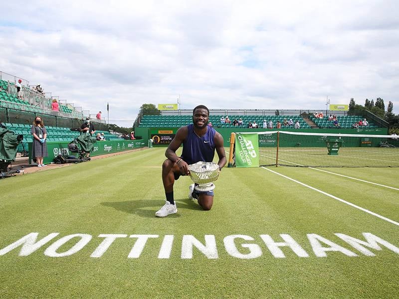 Tennis player smiling and kneeling on the Nottingham grass court while holding a trophy, with 'NOTTINGHAM' written on the court.