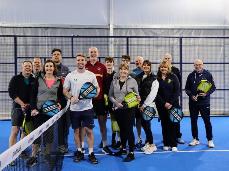 Group of men and women holding padel rackets on a blue indoor padel court, posing in front of the net.