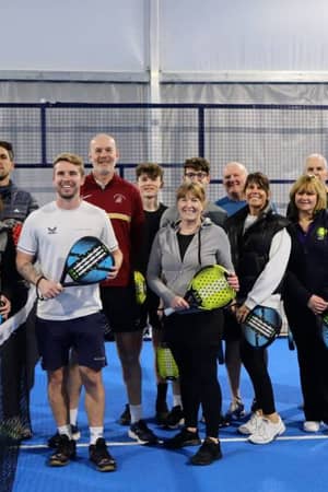 Group of men and women holding padel rackets on a blue indoor padel court, posing in front of the net.