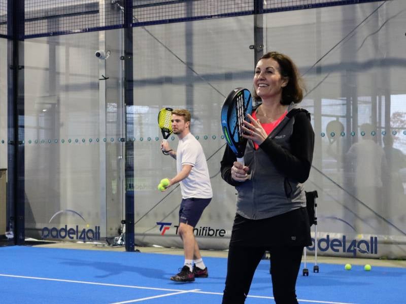 A smiling woman with a padel racket in an indoor court, coaching session under the padel4all brand, man in the background preparing to play.